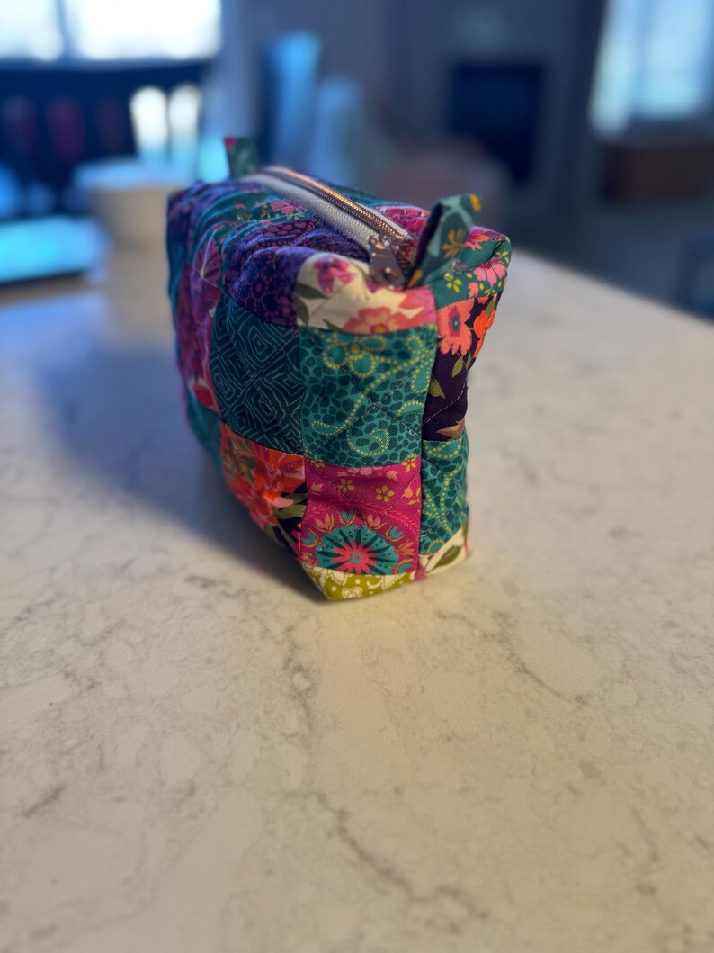 Op de afbeelding: Een rechthoekige, gewatteerde make-up tas met een patchwork ontwerp. De tas heeft verschillende bloemen- en geometrische patronen in tinten van teal, roze, paars en groen. Een zilveren rits loopt over de bovenkant en er zitten kleine stoffen lussen aan elk uiteinde.