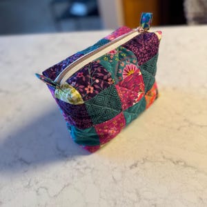 Op de afbeelding: Een gewatteerde make-up tas met een patchwork ontwerp. De tas heeft een verscheidenheid aan stoffen in tinten van teal, paars, roze en groen. Het heeft een ritssluiting met een goudkleurige rits en een kleine stoffen lus aan de zijkant.