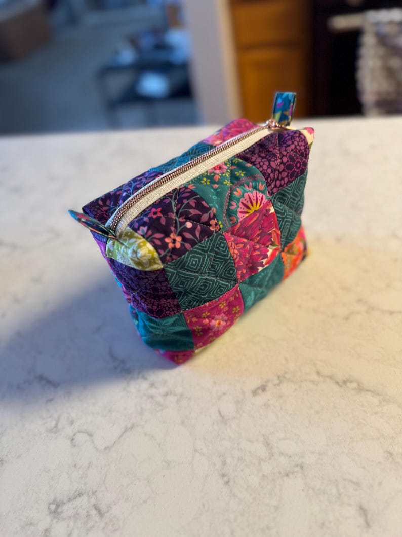 Op de afbeelding: Een gewatteerde make-up tas met een patchwork ontwerp. De tas heeft een verscheidenheid aan stoffen in tinten van teal, paars, roze en groen. Het heeft een ritssluiting met een goudkleurige rits en een kleine stoffen lus aan de zijkant.
