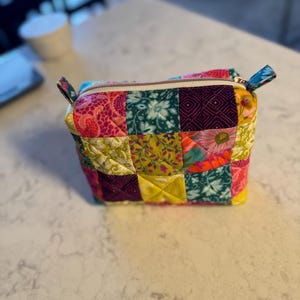 Peut inclure: Petite trousse zipp&eacute;e de style patchwork. Le sac est compos&eacute; de divers tissus &agrave; motifs dans les tons de rose, vert, jaune et turquoise. Il poss&egrave;de une fermeture &eacute;clair blanche et de petites poign&eacute;es turquoise &agrave; motifs.
