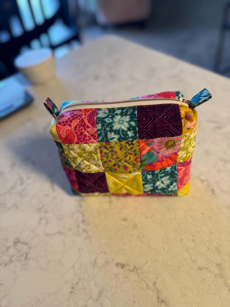 Peut inclure: Petite trousse zipp&eacute;e de style patchwork. Le sac est compos&eacute; de divers tissus &agrave; motifs dans les tons de rose, vert, jaune et turquoise. Il poss&egrave;de une fermeture &eacute;clair blanche et de petites poign&eacute;es turquoise &agrave; motifs.