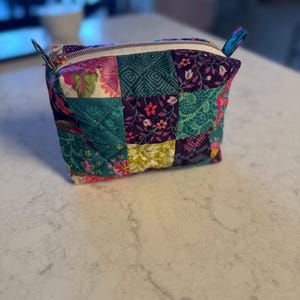 Op de afbeelding: Een gewatteerde patchwork etui met diverse bloemen- en geometrische patronen. De etui heeft stoffen in teal, paars, roze en groen. De witte rits heeft een goudkleurige ritstrekker.