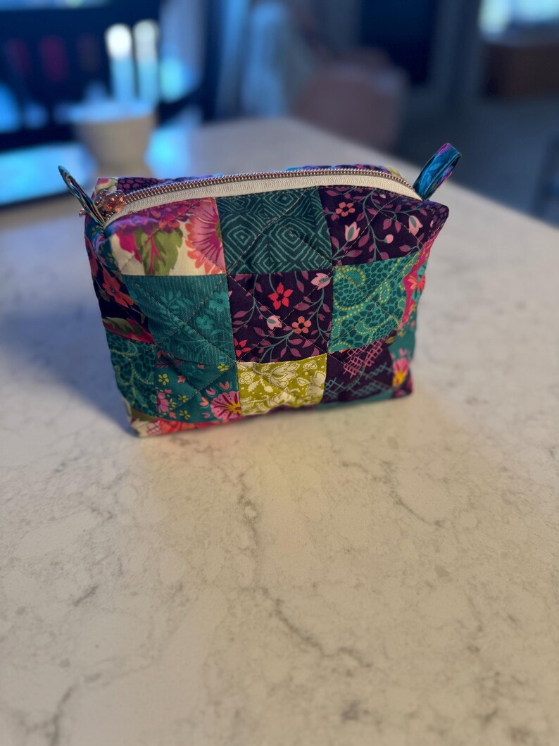 Op de afbeelding: Een gewatteerde patchwork etui met diverse bloemen- en geometrische patronen. De etui heeft stoffen in teal, paars, roze en groen. De witte rits heeft een goudkleurige ritstrekker.