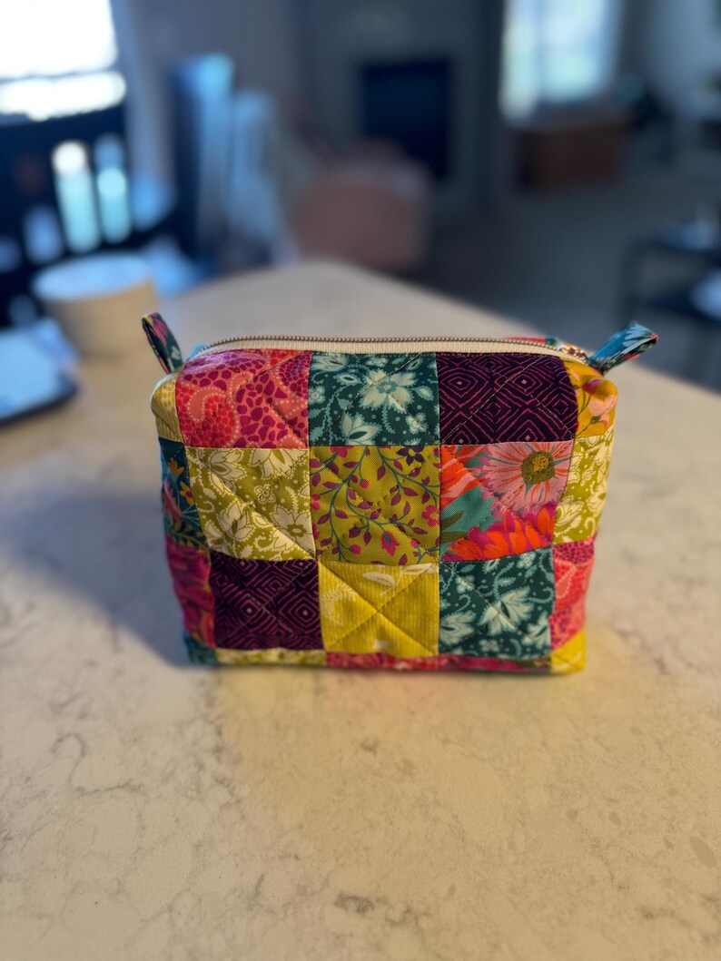 Peut inclure: Petite trousse zipp&eacute;e en patchwork aux motifs floraux et g&eacute;om&eacute;triques multicolores : rose, vert, jaune et violet. Le sac rectangulaire est orn&eacute; de petites boucles en tissu &agrave; chaque extr&eacute;mit&eacute; et d'une fermeture &eacute;clair.