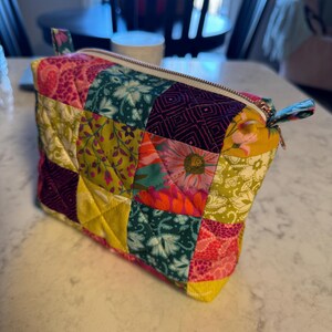 Peut inclure: Une trousse de maquillage matelass&eacute;e avec un motif patchwork. Le sac pr&eacute;sente divers motifs floraux et g&eacute;om&eacute;triques dans les tons de rose, vert, jaune et violet. Il poss&egrave;de une fermeture &eacute;clair et une petite boucle en tissu sur le c&ocirc;t&eacute;.