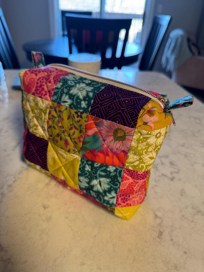Peut inclure: Une trousse de maquillage matelass&eacute;e avec un motif patchwork. Le sac pr&eacute;sente divers motifs floraux et g&eacute;om&eacute;triques dans les tons de rose, vert, jaune et violet. Il poss&egrave;de une fermeture &eacute;clair et une petite boucle en tissu sur le c&ocirc;t&eacute;.