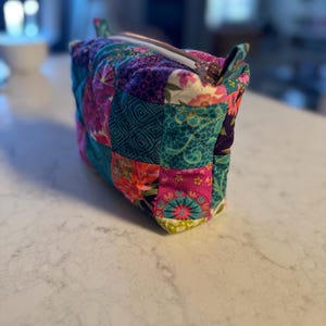 Op de afbeelding: Een kleurrijke, gewatteerde make-uptas met een patchwork-ontwerp. De tas heeft een verscheidenheid aan bloemen- en geometrische patronen in roze, teal, paars en groen. Het heeft een ritssluiting en kleine stoffen handvatten.