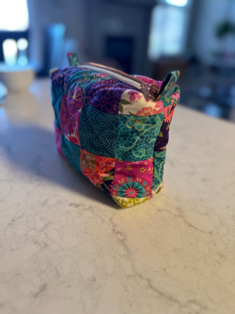 Op de afbeelding: Een kleurrijke, gewatteerde make-uptas met een patchwork-ontwerp. De tas heeft een verscheidenheid aan bloemen- en geometrische patronen in roze, teal, paars en groen. Het heeft een ritssluiting en kleine stoffen handvatten.