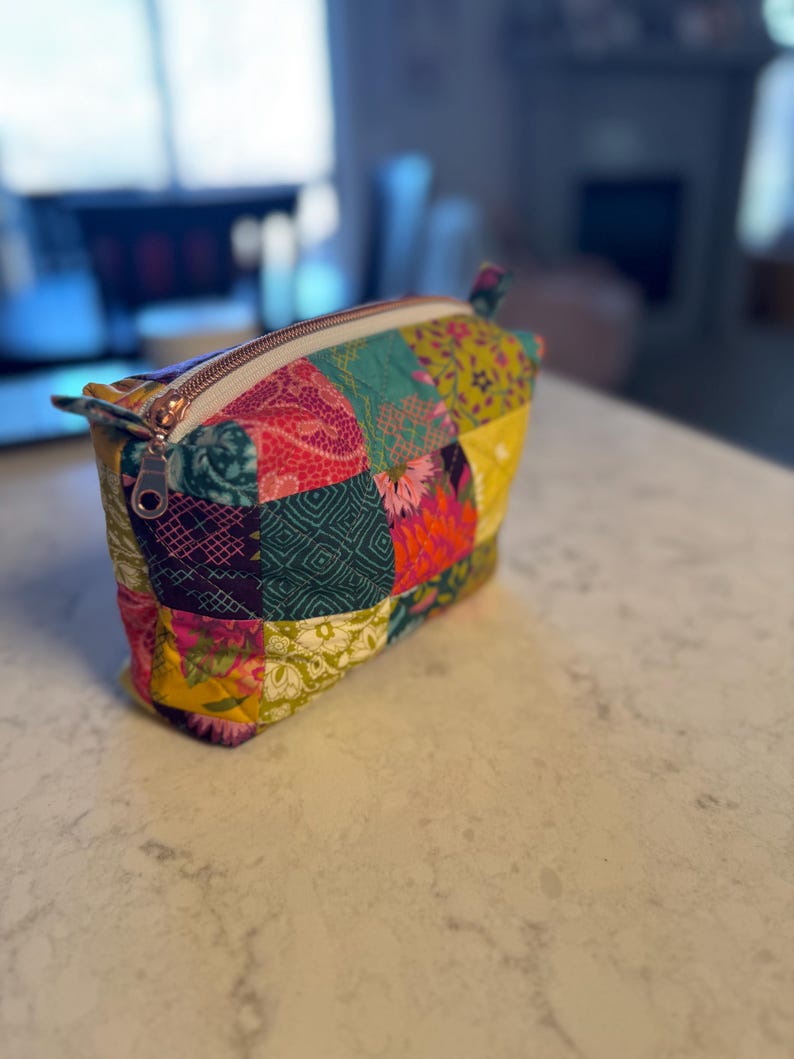 Peut inclure: Une trousse de maquillage patchwork color&eacute;e avec une fermeture &eacute;clair dor&eacute;e. Le sac est compos&eacute; de divers carr&eacute;s de tissu dans les tons de turquoise, rose, jaune et violet. Le sac est rectangulaire et poss&egrave;de une petite boucle sur un c&ocirc;t&eacute;.