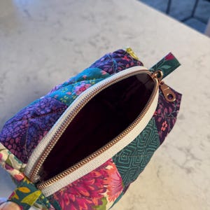 Op de afbeelding: Open, gewatteerd etui met een bloemen patchwork ontwerp in tinten paars, teal en roze. De etui heeft een witte rits met een ros&eacute;gouden trekker en een bijpassend stoffen handvat.