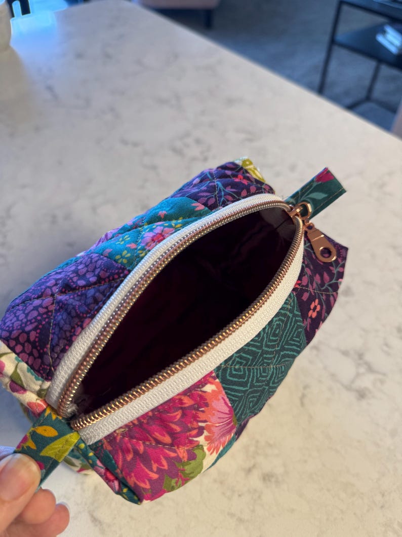 Op de afbeelding: Open, gewatteerd etui met een bloemen patchwork ontwerp in tinten paars, teal en roze. De etui heeft een witte rits met een ros&eacute;gouden trekker en een bijpassend stoffen handvat.