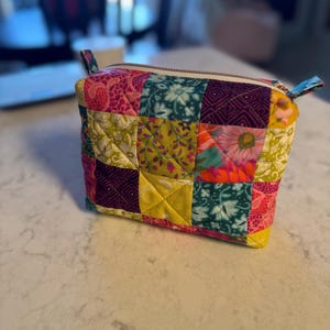 May include: A colorful patchwork zippered pouch with a variety of floral and geometric patterns in pink, green, yellow, and teal. The pouch has a white zipper and small fabric loops on each end.