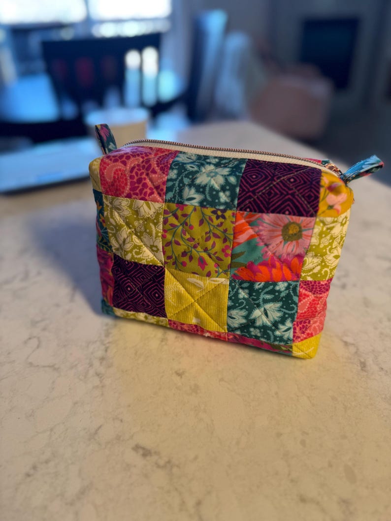 Peut inclure: Une trousse &agrave; fermeture &eacute;clair en patchwork color&eacute;, avec divers motifs floraux et g&eacute;om&eacute;triques en rose, vert, jaune et turquoise. La trousse a une fermeture &eacute;clair blanche et des boucles en tissu.