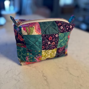 Op de afbeelding: Een gewatteerde make-uptas met een patchwork-ontwerp. De tas heeft een verscheidenheid aan stoffen in tinten van teal, paars, roze en groen. Het heeft een witte rits en kleine stoffen lussen aan elke kant. De tas staat op een wit aanrecht.