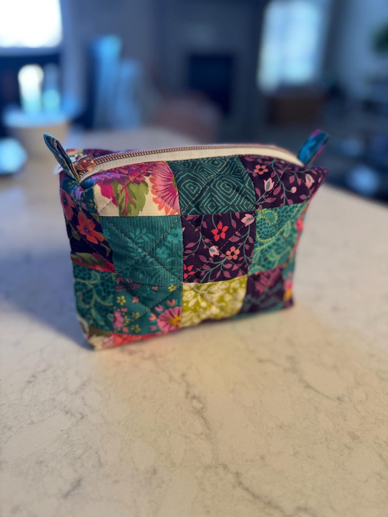 Op de afbeelding: Een gewatteerde make-uptas met een patchwork-ontwerp. De tas heeft een verscheidenheid aan stoffen in tinten van teal, paars, roze en groen. Het heeft een witte rits en kleine stoffen lussen aan elke kant. De tas staat op een wit aanrecht.