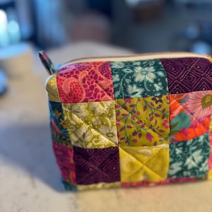 Peut inclure: Un sac &agrave; cosm&eacute;tiques matelass&eacute; aux motifs patchwork. Il pr&eacute;sente des carr&eacute;s aux motifs floraux et g&eacute;om&eacute;triques vari&eacute;s dans les tons rose, vert, jaune et turquoise. Il est dot&eacute; d'une fermeture &eacute;clair blanche et d'une petite boucle en tissu. Le sac est pos&eacute; sur une surface blanche.