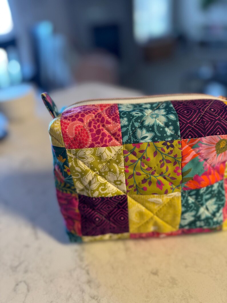 Peut inclure: Un sac &agrave; cosm&eacute;tiques matelass&eacute; aux motifs patchwork. Il pr&eacute;sente des carr&eacute;s aux motifs floraux et g&eacute;om&eacute;triques vari&eacute;s dans les tons rose, vert, jaune et turquoise. Il est dot&eacute; d'une fermeture &eacute;clair blanche et d'une petite boucle en tissu. Le sac est pos&eacute; sur une surface blanche.