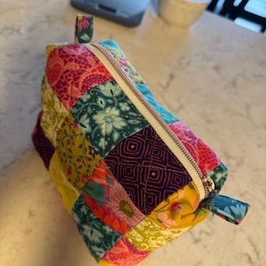 Peut inclure: Une trousse zipp&eacute;e en patchwork color&eacute;, orn&eacute;e de motifs floraux et g&eacute;om&eacute;triques dans des tons de rose, jaune, turquoise et violet. La trousse est dot&eacute;e d'une fermeture &eacute;clair dor&eacute;e et de petites boucles en tissu &agrave; chaque extr&eacute;mit&eacute;.