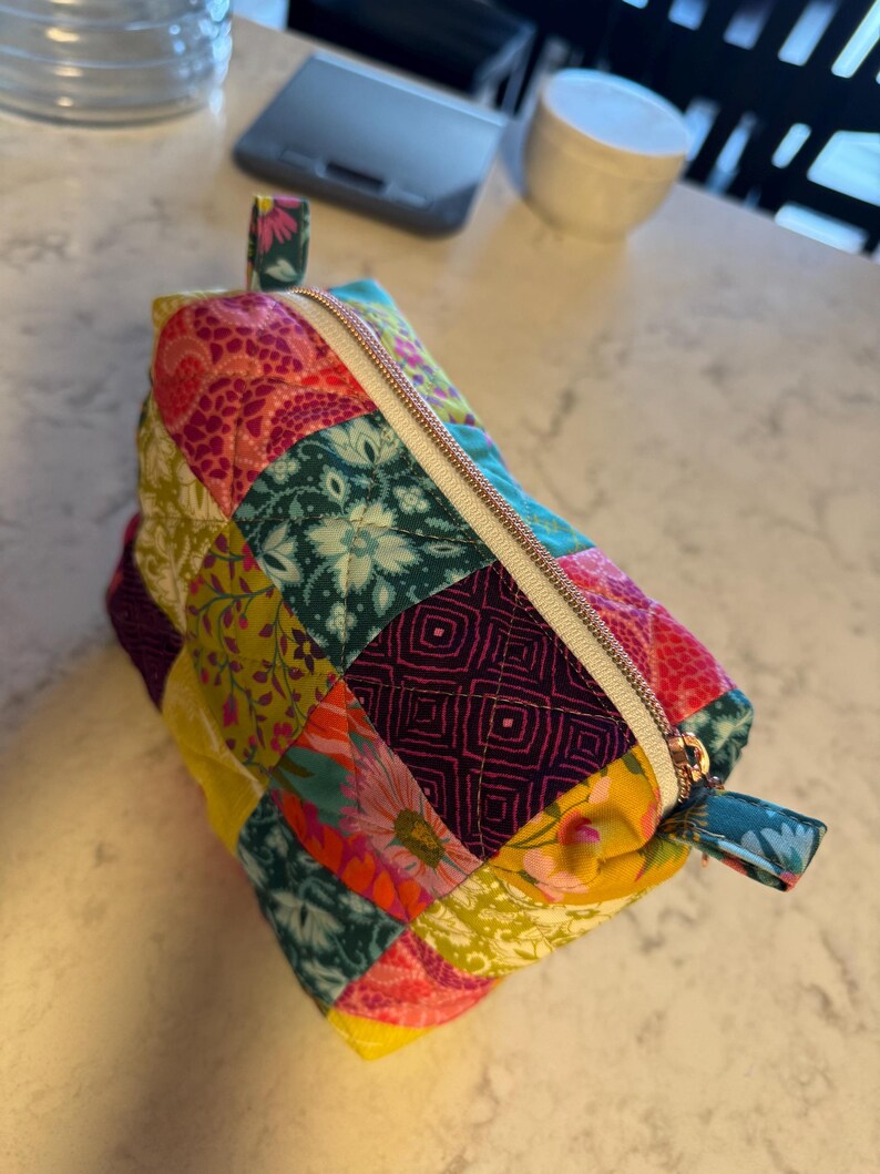 Peut inclure: Une trousse zipp&eacute;e en patchwork color&eacute;, orn&eacute;e de motifs floraux et g&eacute;om&eacute;triques dans des tons de rose, jaune, turquoise et violet. La trousse est dot&eacute;e d'une fermeture &eacute;clair dor&eacute;e et de petites boucles en tissu &agrave; chaque extr&eacute;mit&eacute;.