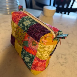 Peut inclure: Une trousse de maquillage patchwork color&eacute;e avec une fermeture &eacute;clair dor&eacute;e. Le sac est fait de divers tissus &agrave; motifs dans les tons de rose, jaune, vert et violet. Il a une petite boucle en tissu.