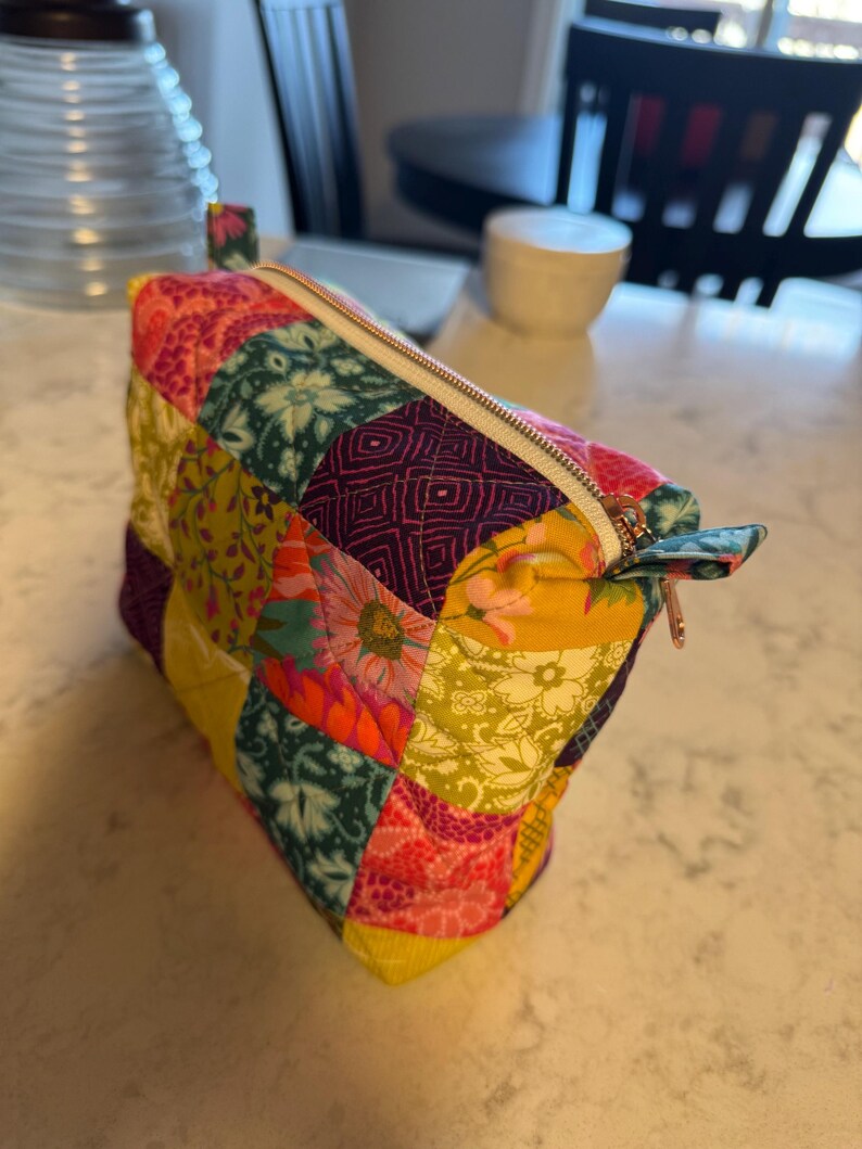 Peut inclure: Une trousse de maquillage patchwork color&eacute;e avec une fermeture &eacute;clair dor&eacute;e. Le sac est fait de divers tissus &agrave; motifs dans les tons de rose, jaune, vert et violet. Il a une petite boucle en tissu.