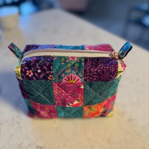 Op de afbeelding: Een gewatteerde make-up tas met een patchwork ontwerp. De tas heeft een verscheidenheid aan kleurrijke bloemen- en geometrische patronen in tinten paars, teal, roze en oranje. Het heeft een ritssluiting met een goudkleurige trekker en kleine stoffen lussen.