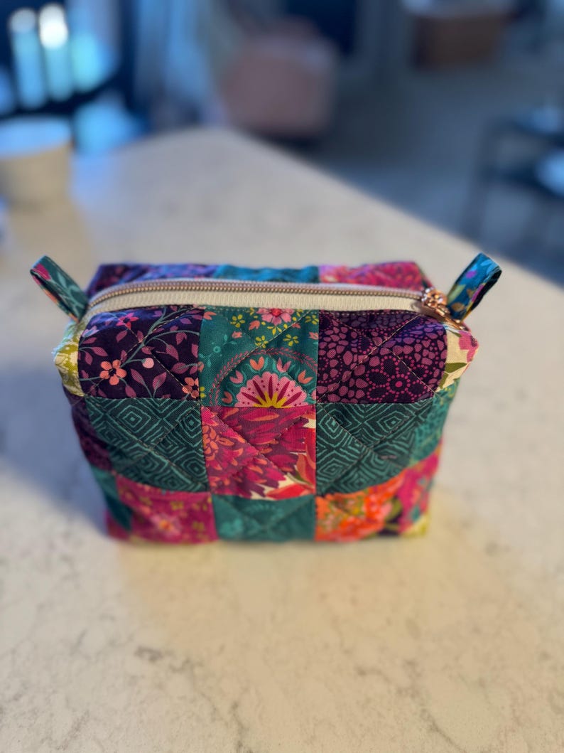 Op de afbeelding: Een gewatteerde make-up tas met een patchwork ontwerp. De tas heeft een verscheidenheid aan kleurrijke bloemen- en geometrische patronen in tinten paars, teal, roze en oranje. Het heeft een ritssluiting met een goudkleurige trekker en kleine stoffen lussen.
