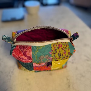 Peut inclure: Trousse de maquillage patchwork ouverte avec une fermeture &eacute;clair dor&eacute;e. Le sac pr&eacute;sente une vari&eacute;t&eacute; de motifs floraux et g&eacute;om&eacute;triques dans des tons de rose, vert, jaune et violet. L'int&eacute;rieur est de couleur magenta fonc&eacute;.