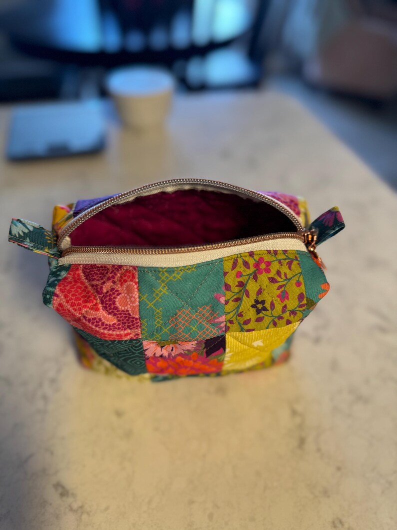 Peut inclure: Trousse de maquillage patchwork ouverte avec une fermeture &eacute;clair dor&eacute;e. Le sac pr&eacute;sente une vari&eacute;t&eacute; de motifs floraux et g&eacute;om&eacute;triques dans des tons de rose, vert, jaune et violet. L'int&eacute;rieur est de couleur magenta fonc&eacute;.
