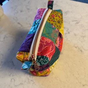 Peut inclure: Une trousse de maquillage matelass&eacute;e color&eacute;e avec un motif patchwork. Le sac pr&eacute;sente une vari&eacute;t&eacute; de tissus dans les tons de violet, rose, jaune et sarcelle. Une fermeture &eacute;clair en or rose longe le dessus. Le sac a une petite boucle en tissu &agrave; une extr&eacute;mit&eacute;.