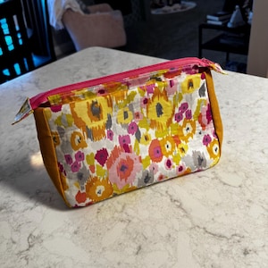 May include: A rectangular cosmetic bag with a floral pattern in yellow, orange, pink, and gray. The bag has a bright pink zipper and mustard-colored sides. The bag is sitting on a white marble countertop.