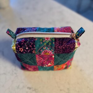 Op de afbeelding: Een patchwork cosmetische tas met een rechthoekige vorm. De tas is gemaakt van gewatteerde stof met bloemen- en geometrische patronen in tinten van teal, paars, roze en geel. Het heeft een gouden rits en kleine stoffen lussen aan elk uiteinde.