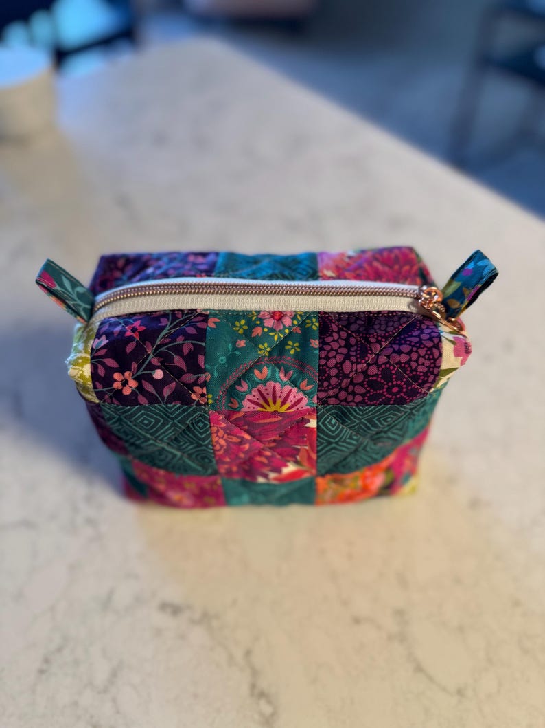 Op de afbeelding: Een patchwork cosmetische tas met een rechthoekige vorm. De tas is gemaakt van gewatteerde stof met bloemen- en geometrische patronen in tinten van teal, paars, roze en geel. Het heeft een gouden rits en kleine stoffen lussen aan elk uiteinde.