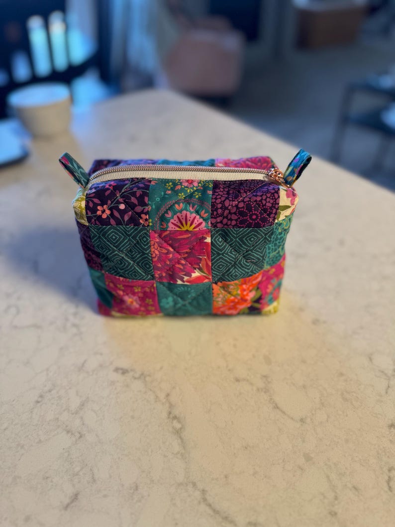 Op de afbeelding: Een kleine, gewatteerde etui met een patchwork-ontwerp. De etui heeft een verscheidenheid aan kleurrijke stoffen in tinten roze, paars, blauwgroen en oranje. Het heeft een ritssluiting met een goudkleurige rits en kleine stoffen lussen aan elke kant.