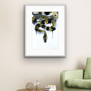 Könnte beinhalten: Gerahmtes Aquarellgemälde einer Schlange in Schwarz, Gold und Weiß. Die Schlange ist stilisiert und abstrakt dargestellt, mit schwarzen Farbtropfen. Das Kunstwerk wird auf einem weißen Passepartout in einem silbernen Rahmen präsentiert.