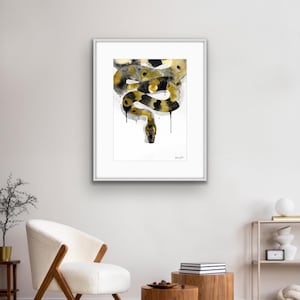 Könnte beinhalten: Ein gerahmter Aquarelldruck einer Schlange in Schwarz-, Gold- und Grautönen. Die Schlange ist in einem stilisierten, abstrakten Stil dargestellt, wobei der Kopf nach unten zeigt. Das Kunstwerk wird in einem weißen Rahmen präsentiert und verleiht dem Raum eine moderne Note.