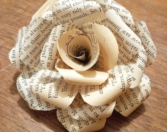 Puede incluir: Una rosa de papel hecha con páginas de libros. Los pétalos superpuestos revelan texto, creando un efecto literario único. La flor, de color crema, está colocada sobre una superficie de madera.