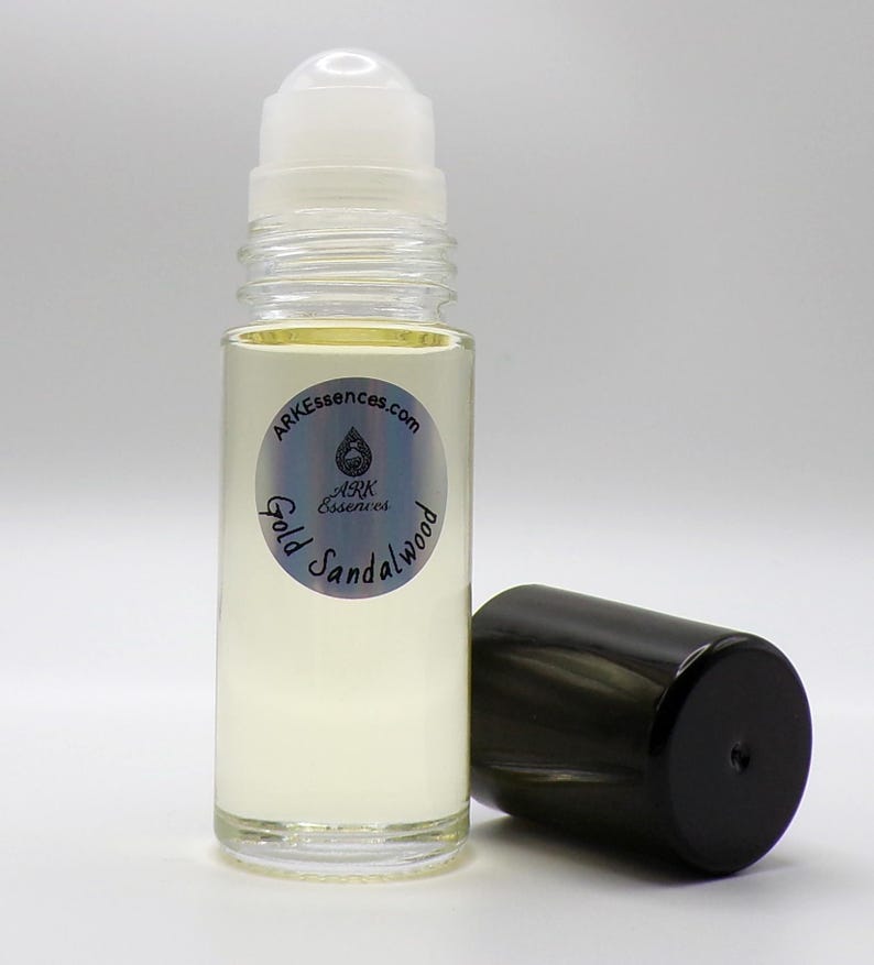May include: A clear glass roll-on bottle containing a pale yellow liquid. The bottle has a white rollerball top and a black cap. The label reads "Gold Sandalwood" and "ARKessences.com".