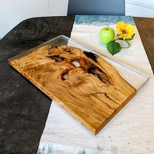 May include: A rectangular wooden serving board with a live edge and a clear resin inlay. The wood has a light brown color with dark brown and black patterns. The resin section is a light gray color.