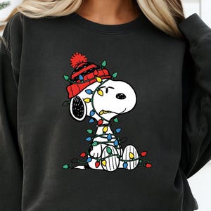 May include: Black sweatshirt featuring a graphic of Snoopy wearing a red and black hat and wrapped in Christmas lights. The lights are multi-colored. Snoopy is a white dog with black ears and a black nose.