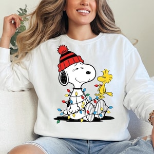 May include: White sweatshirt featuring Snoopy wearing a red and black knit cap and a string of colorful Christmas lights. Woodstock is perched beside Snoopy. The design is a festive holiday theme.