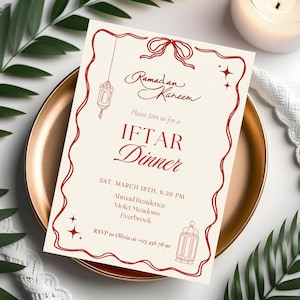 May include: An invitation for an Iftar dinner, featuring the text "Ramadan Kareem" and "IFTAR Dinner" in red script. The invitation is on a cream background with a red border and decorative lanterns. It is placed on a gold plate with greenery.