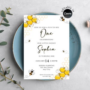 以下が含まれることがあります： 「One Celebration! Our Little Honey Sophia is turning one」のテキストが特徴の、ミツバチをテーマにした白い招待状。カードにはミツバチのイラストとハニカム模様が含まれています。日付と時間も含まれています。