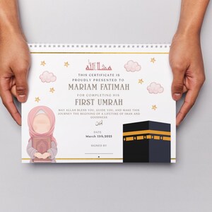 May include: A certificate with a spiral binding, held by hands. The certificate features text in English and Arabic, with illustrations of stars, clouds, and a Kaaba. The certificate is presented to Mariam Fatimah for completing her First Umrah.