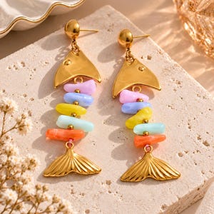 Peut inclure: Une paire de boucles d'oreilles pendantes en forme de poisson doré avec une tête et une queue en métal poli. Le corps de chaque poisson est composé de cinq perles lisses et multicolores dans des tons lavande, bleu clair, jaune, turquoise et orange.