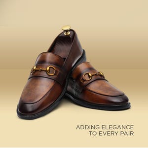Puede incluir: Mocasines de cuero marrón con detalles dorados de herradura. Los zapatos tienen un acabado pulido y un diseño clásico. El texto "ADDING ELEGANCE TO EVERY PAIR" es visible en la parte inferior.