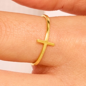May include: A gold-colored ring featuring a simple cross design. The ring is worn on a finger, showcasing its minimalist style. The cross is a prominent, central element, adding a touch of faith and elegance to the jewelry.