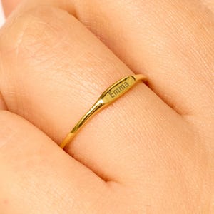 May include: A gold-colored ring with the name "Emma" engraved on it. The ring is thin and worn on a finger. The background is a light peach color.