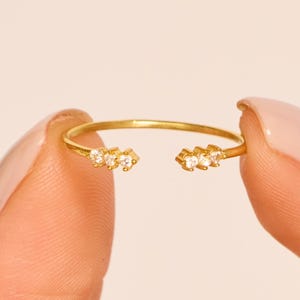 May include: Open-ended gold ring with three small, clear gemstones on each end. The ring is held between two fingers against a soft, neutral background. The ring's design is minimalist and elegant.