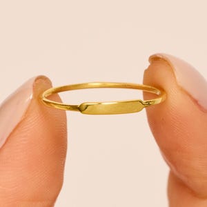Könnte beinhalten: Ein zarter goldener Ring mit einem rechteckigen Mittelteil. Der Ring wird zwischen zwei Fingern vor einem weichen, neutralen Hintergrund gehalten. Das Design des Rings ist schlicht und elegant, geeignet für den Alltag.